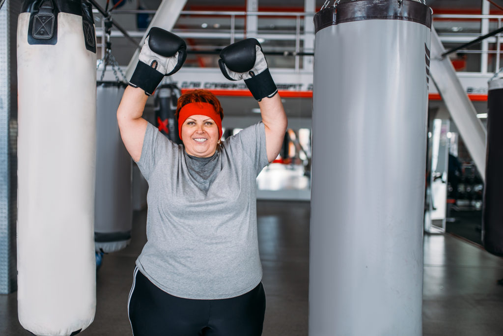 Overweight sweaty woman in boxing gloves against punching bag in gym. Calories burning, obese female person on hard training in sport club Weight loss and nutrition Complete Healthcare Primary Care and Gynecology