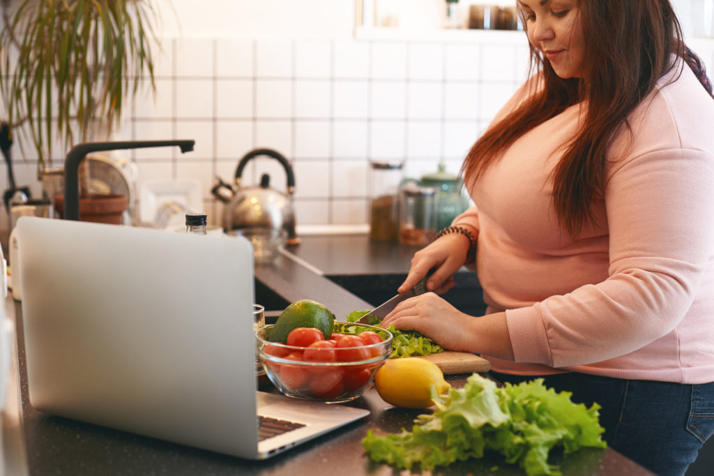 Overweight woman using laptop to watch video recipe while making vegan vitamin avocado salad, slicing leaf lettuce on wooden cutting board. Healthy food, weight loss, dieting and nutrition concept Complete Healthcare Primary Care and Gynecology