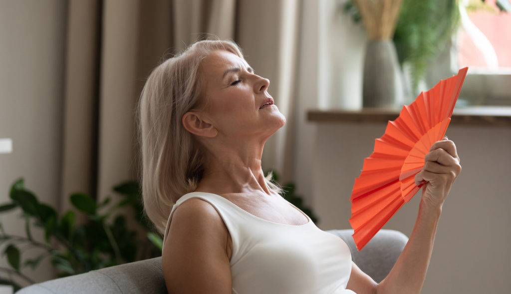 Older woman having a hot flash on her couch from menopause Older woman having a hot flash from menopause and using a fan Complete Healthcare Primary Care and Gynecology