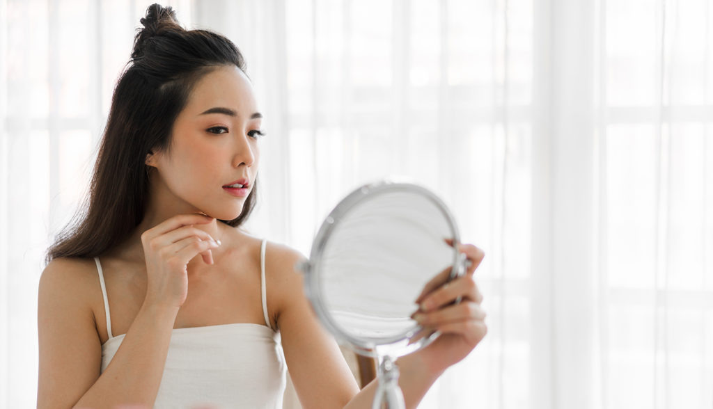 Young Asian woman holding mirror and looking at herself and inspecting her chin on white background Complete Healthcare Primary Care and Gynecology