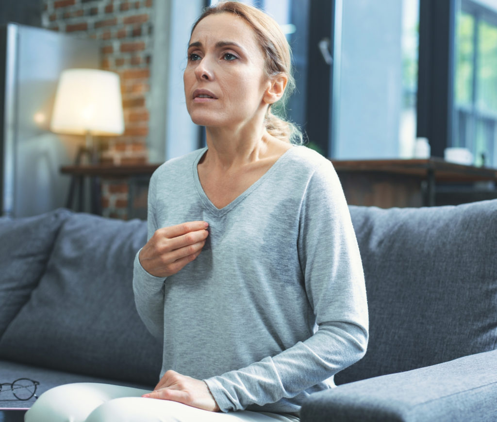 Older woman having a hot flash sitting on her couch in the living room Complete Healthcare Primary Care and Gynecology