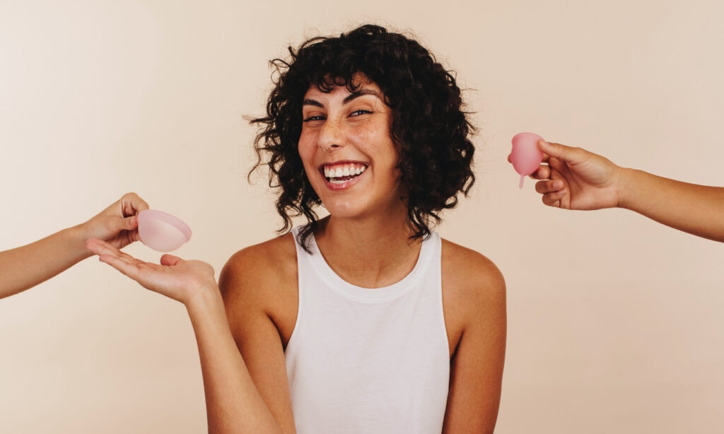 Smiling woman with short, curlt black hair wearing a white tank top being handed a menstrual cup and menstrual discs by two hands offscreen
