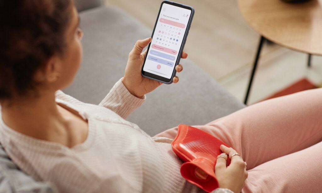 Woman sitting and holding a heating pad against her uterus while using a period tracker app